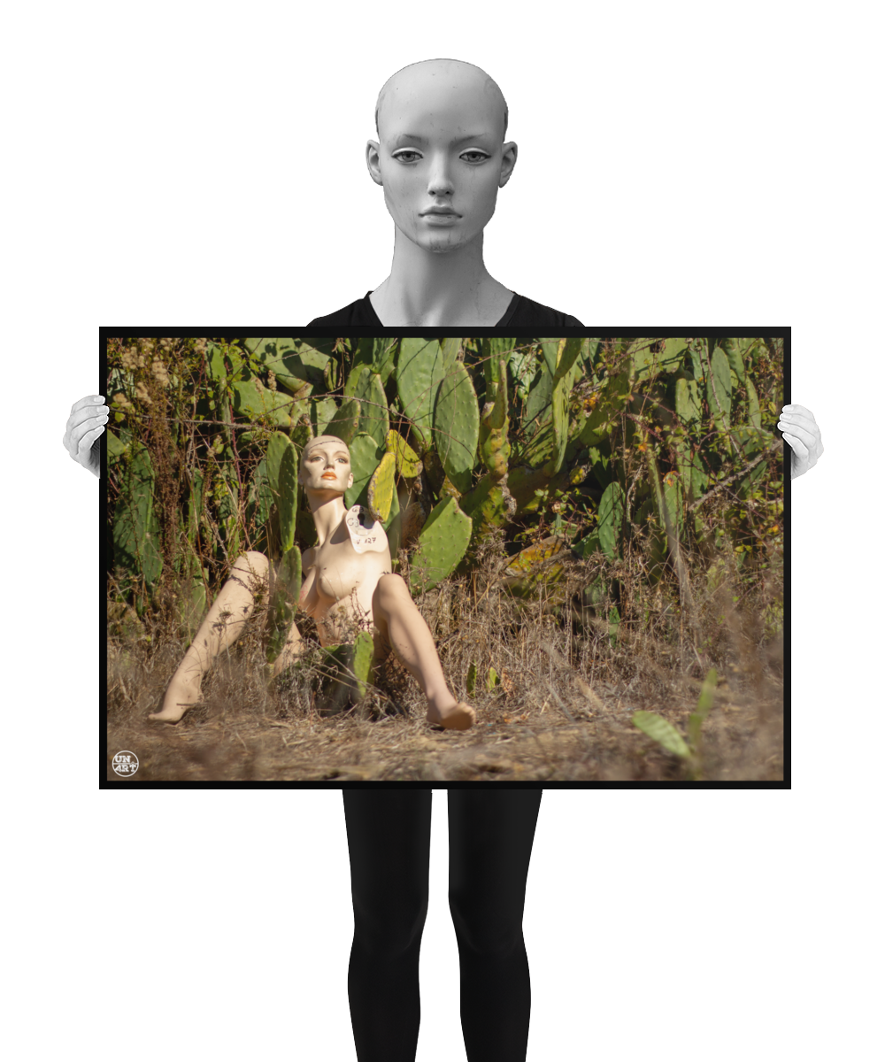 un4rt - almost human gallery poster mockup picture with a mannequin sitting outdoor in in front of a huge cactus. dried gras on the ground. the poster is held by a mannequin.