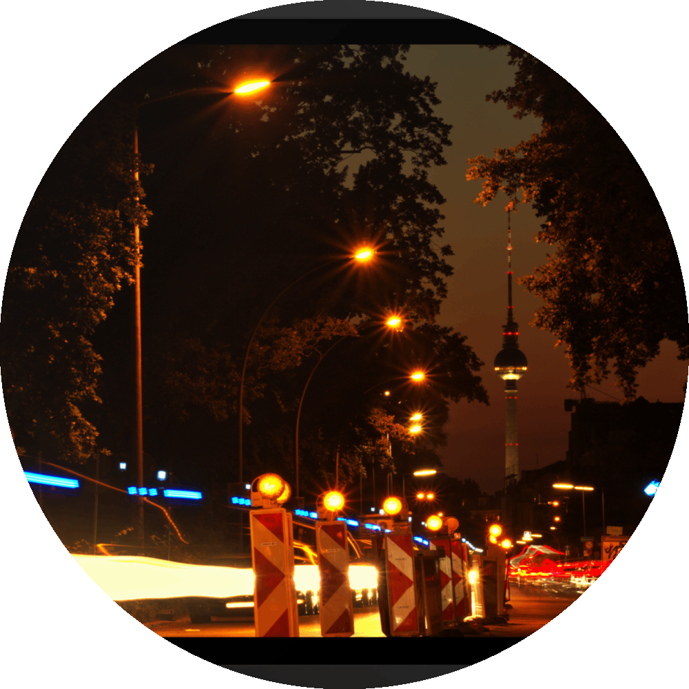 un4rt - the photographie shows a street in berlin. the street is called "puschkinallee" and the view goes along a construction site. at the end of the street berlin´s television tower is clearly visible. time lapse. golden hour. trees and city lights.