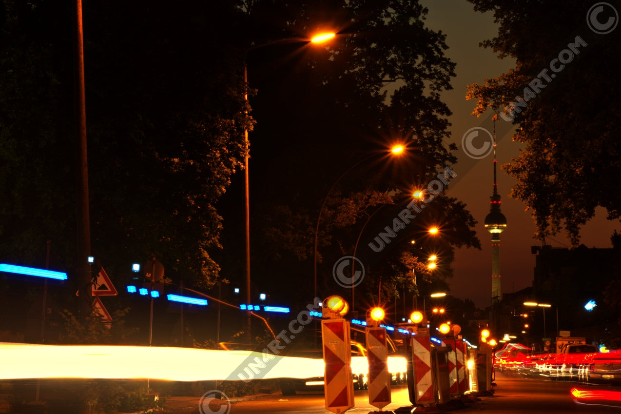 un4rt - the photographie shows a street in berlin. the street is called "puschkinallee" and the view goes along a construction site. at the end of the street berlin´s television tower is clearly visible. time lapse. golden hour. trees and city lights.