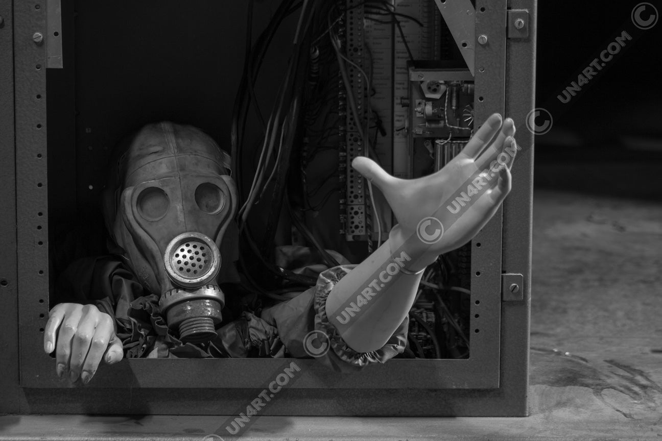 un4rt - black and white photographie of a mannequin wearing a gas mask. the mannequin stretches out one arm and reaches for help. the body is stuck inside a box with wires and electronic parts.