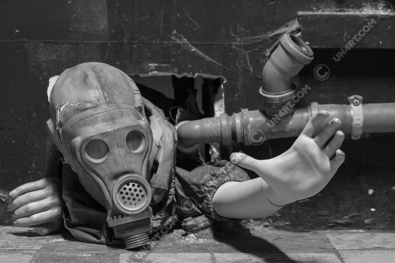 un4rt - black and white photographie of a mannequin wearing a gas mask. the mannequin stretches out one arm and reaches for help. the body is stuck inside a small hole in a wall. unused blackwater pipes next to the mannequin.