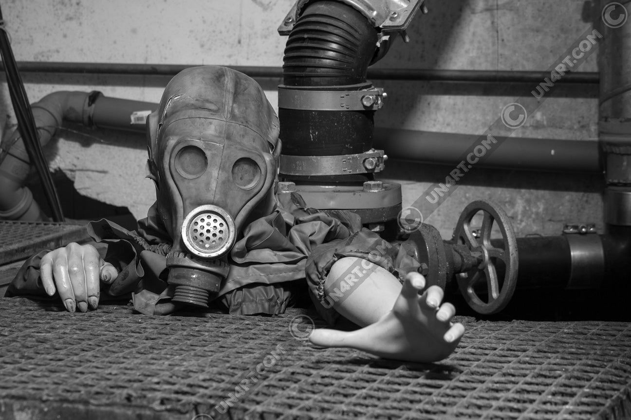 un4rt - black and white photographie of a mannequin wearing a gas mask. the mannequin stretches out one arm and reaches for help. the mannequin is crawling out of a hole in the ground. grif floor ground. pipes and a shut-off valve next to the mannequin.