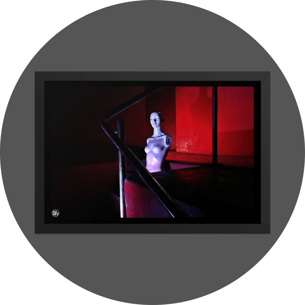 un4rt - the photographie shows the naked upper body of a mannequin without arms in the middle of a staircase. the mannequin is bold. the walls are all red, but due to the minimalistic lighting everything - except the mannequin - looks very dark. the point of view goes through a steel pipe handrail.