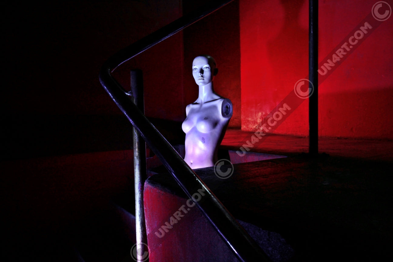 un4rt - the photographie shows the naked upper body of a mannequin without arms in the middle of a staircase. the mannequin is bold. the walls are all red, but due to the minimalistic lighting everything - except the mannequin - looks very dark. the point of view goes through a steel pipe handrail.