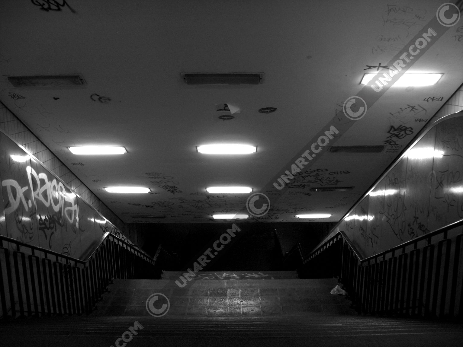 un4rt - a black and white photographie of a former underground trespas that once was part of the train station "alexanderplatz in berlin. a stairway that is leading down. graffities on the ceiling and the wall.