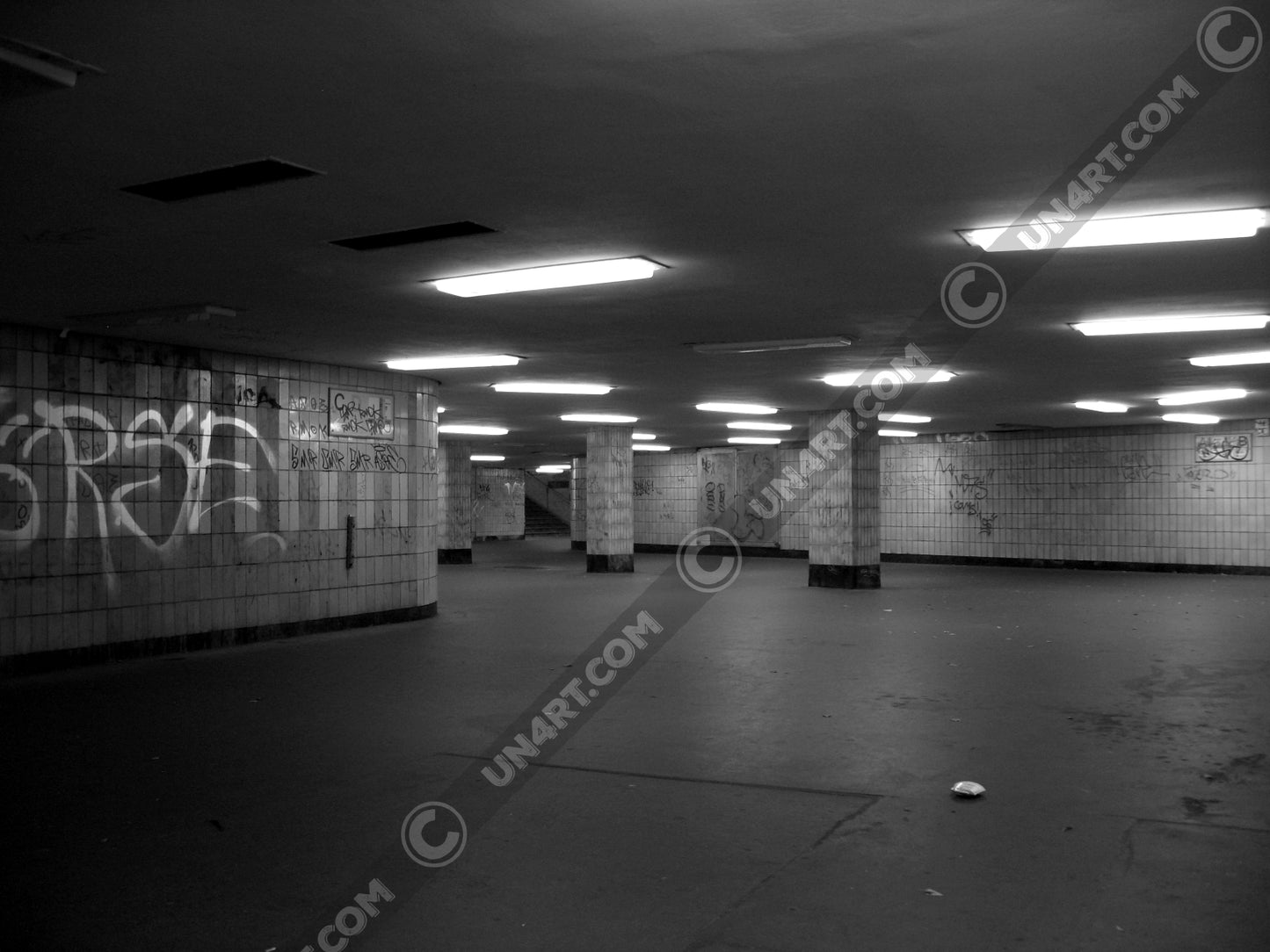 un4rt - a black and white photographie of a former underground trespas that once was part of the train station "alexanderplatz in berlin. graffities on the wall, trash on the ground. some lights on the ceiling don´t work. a stairway at the end of the trespas.