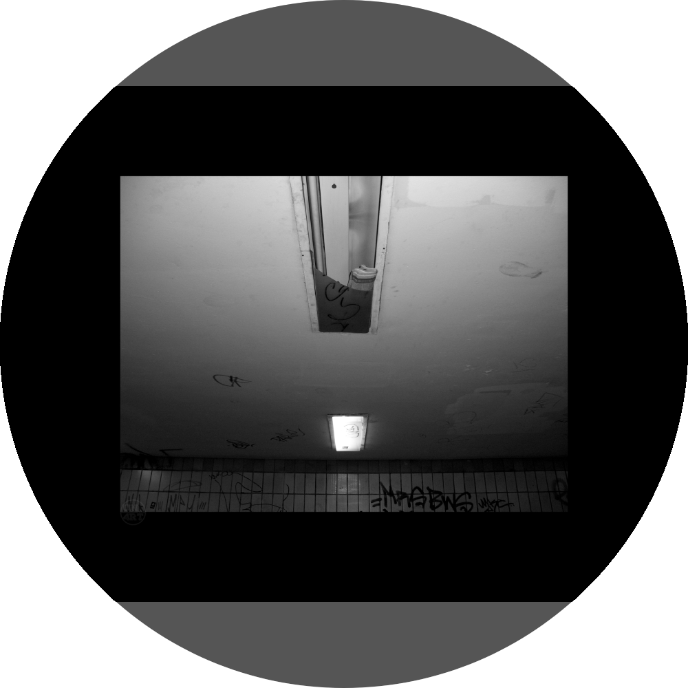 un4rt - a black and white photographie of a former underground trespas that once was part of the train station "alexanderplatz in berlin. the wall is tiled and coverd with graffities. one of the two lights on the ceiling don´t work anymore. the glas is broken and a newspaper is stuffed into the lamp.