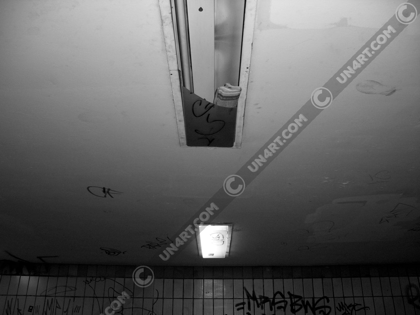 un4rt - a black and white photographie of a former underground trespas that once was part of the train station "alexanderplatz in berlin. the wall is tiled and coverd with graffities. one of the two lights on the ceiling don´t work anymore. the glas is broken and a newspaper is stuffed into the lamp.