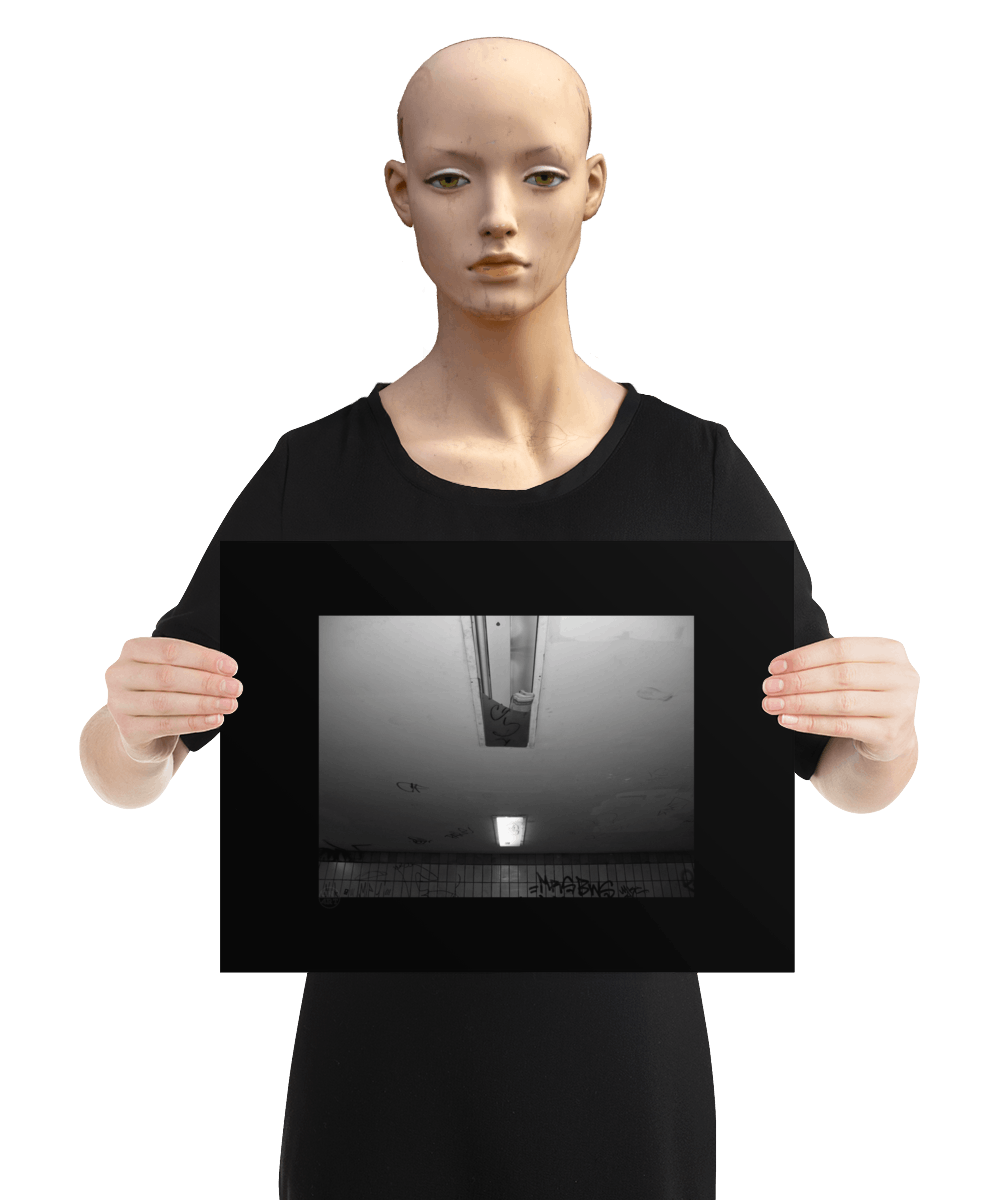 un4rt - a black and white photographie of a former underground trespas that once was part of the train station "alexanderplatz in berlin. the wall is tiled and coverd with graffities. one of the two lights on the ceiling don´t work anymore. the glas is broken and a newspaper is stuffed into the lamp. the image is held by a mannequin.