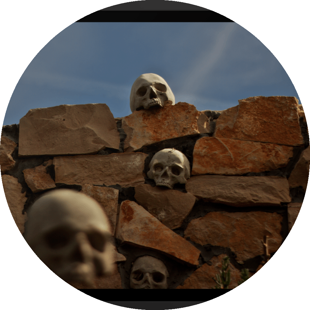 un4rt - the photographie shows a chaotic decorative stone wall with skulls in and on top of it. chemtrails in the sky starting to cover the natural blue of the sky.. one blurry skull in the foregrund.