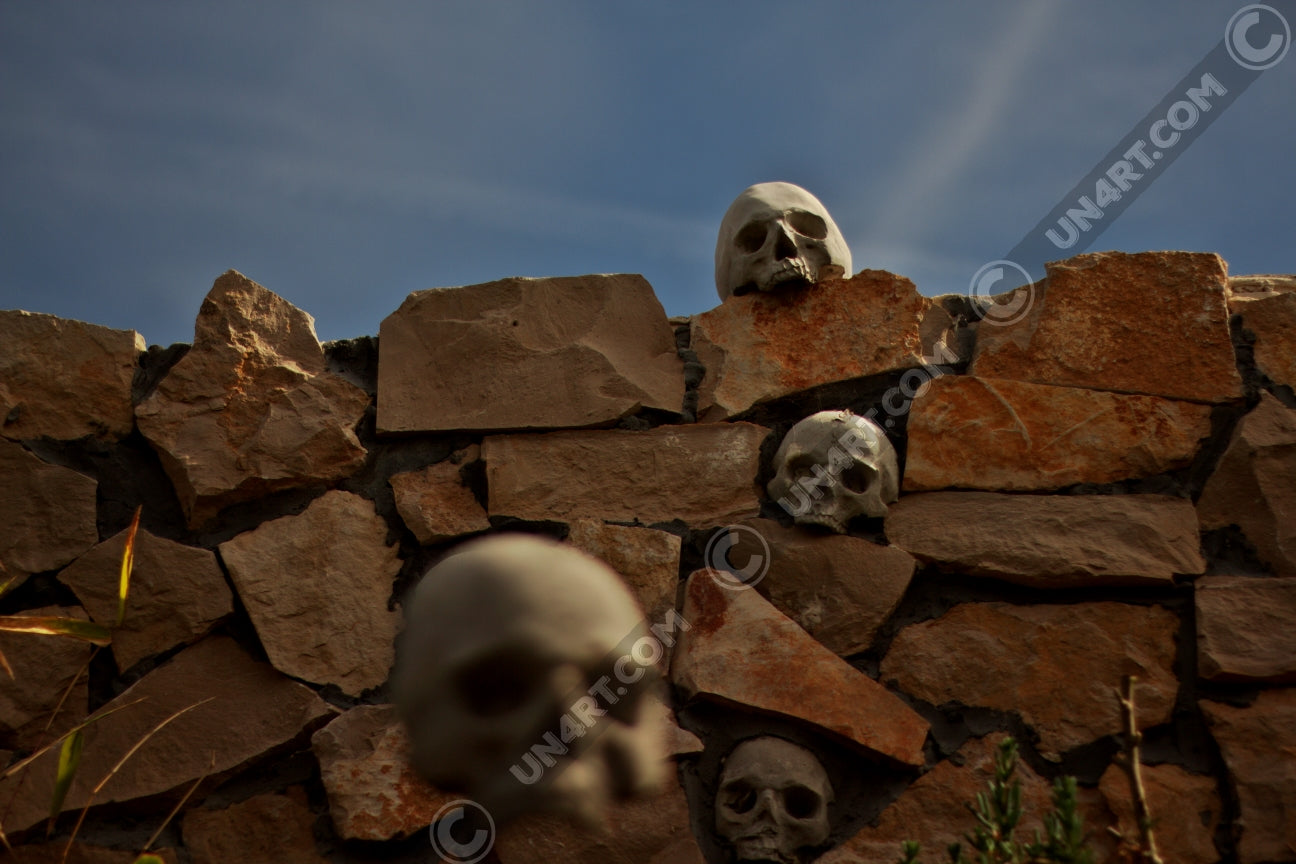 un4rt - the photographie shows a chaotic decorative stone wall with skulls in and on top of it. chemtrails in the sky starting to cover the natural blue of the sky.. one blurry skull in the foregrund.