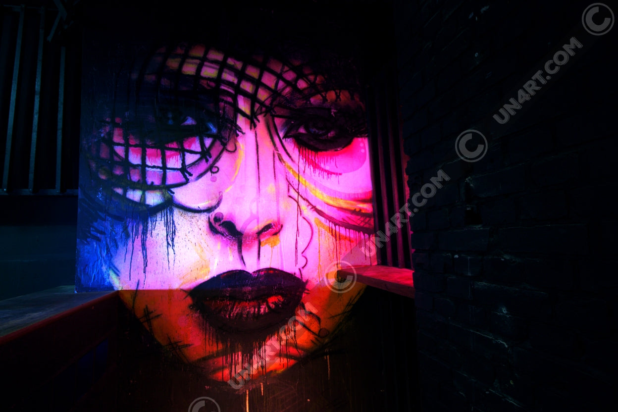 un4rt - the photographie shows a mural of a female face in various colors, pink, yellow, blue, orange. the face is surrounded by black walls, some made of bricks, some made of wood.