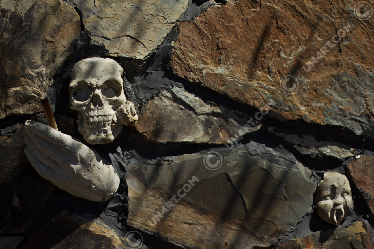 un4rt - the photographie shows some stones that are art of a bigger stone wall. a plaster skull with a rose in its mouth, a plaster hand with a joint and a baby face with plaster vampire teeth decorate the wall.