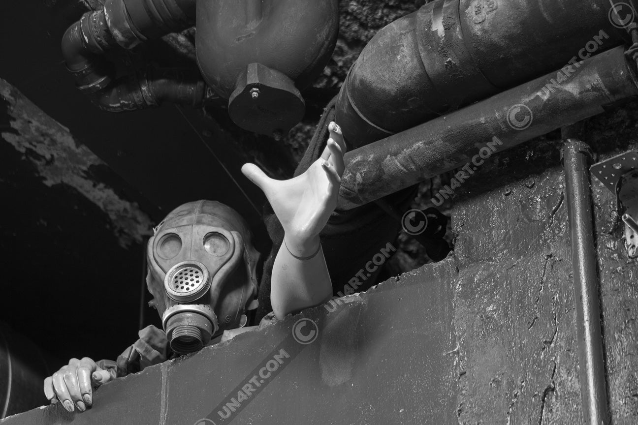 un4rt - black and white photographie of a mannequin wearing a gas mask. the mannequin stretches out one arm and reaches for help. the mannequin is stuck between a wall and a ceiling inside a cellar. waste water pipes above the mannequin.