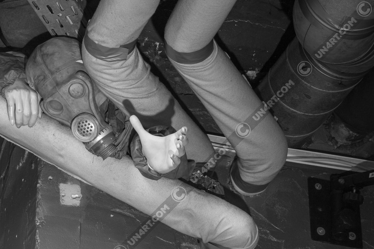 un4rt - black and white photographie of a mannequin wearing a gas mask. the mannequin stretches out one arm and reaches for help. the mannequin is stuck between pipes under the ceiling inside a cellar. waste water pipes around the mannequin.