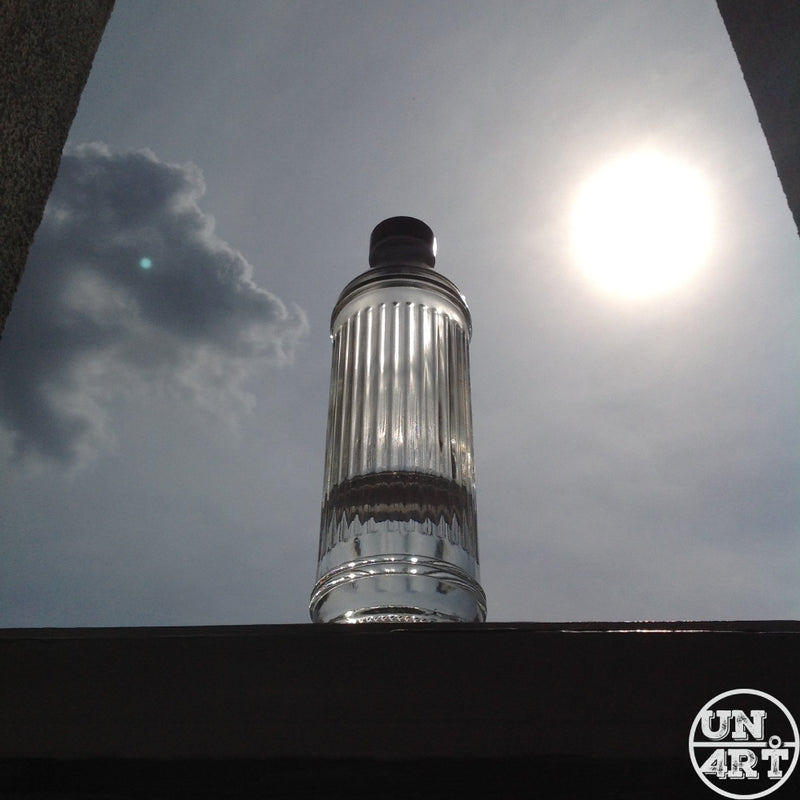 UN4RT imaginarium – ribbed glass bottle silhouetted against a bright sun and overcast sky, low-angle minimalist outdoor photography.