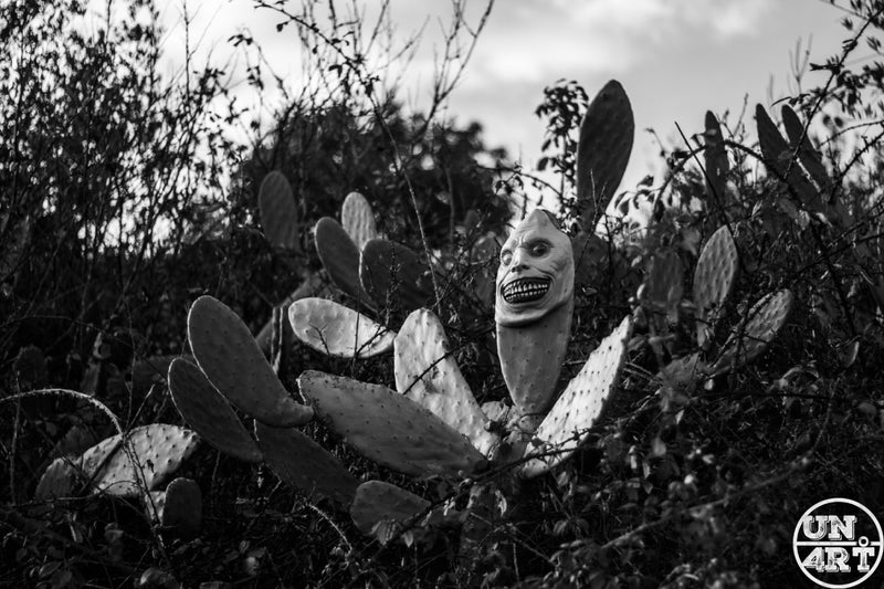 UN4RT imaginarium – grotesque grinning alien mask emerging from a prickly pear cactus thicket, black and white horror photography.