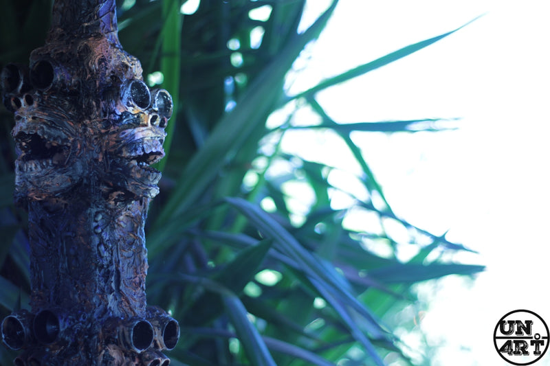 UN4RT imaginarium – dark blue textured tree trunk with eroded skull-like cavities and hollow eye sockets emerging from dense tropical foliage in soft backlight, dark uncanny nature photography.