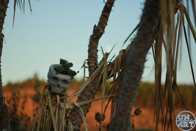 UN4RT imaginarium – weathered concrete skull wedged between dry palm trunks with drooping dead fronds against a warm blue desert sky at golden hour, dark anthropological outdoor art photography.