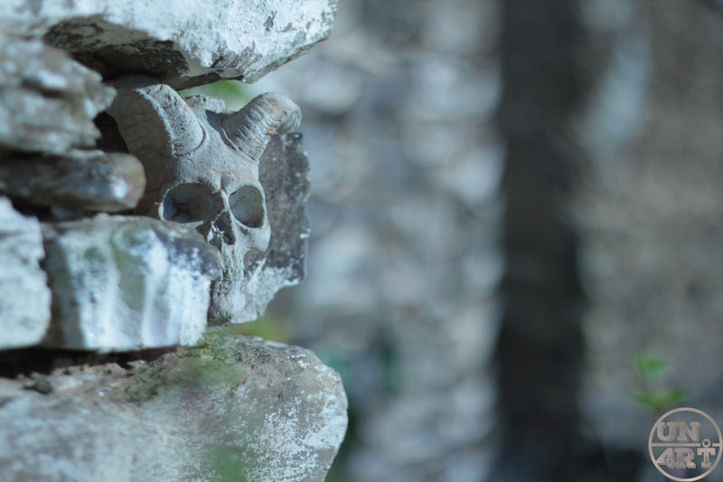 UN4RT imaginarium – small weathered stone skull wedged between stacked mossy limestone rocks with shallow depth of field blurring the surrounding stonework, dark uncanny nature macro photography.