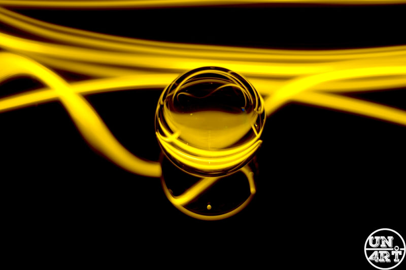 UN4RT imaginarium – small transparent glass marble reflecting golden neon light streaks resting on a mirrored black surface with sweeping amber light trails in the background, dark minimalist fine art light painting macro photography.
