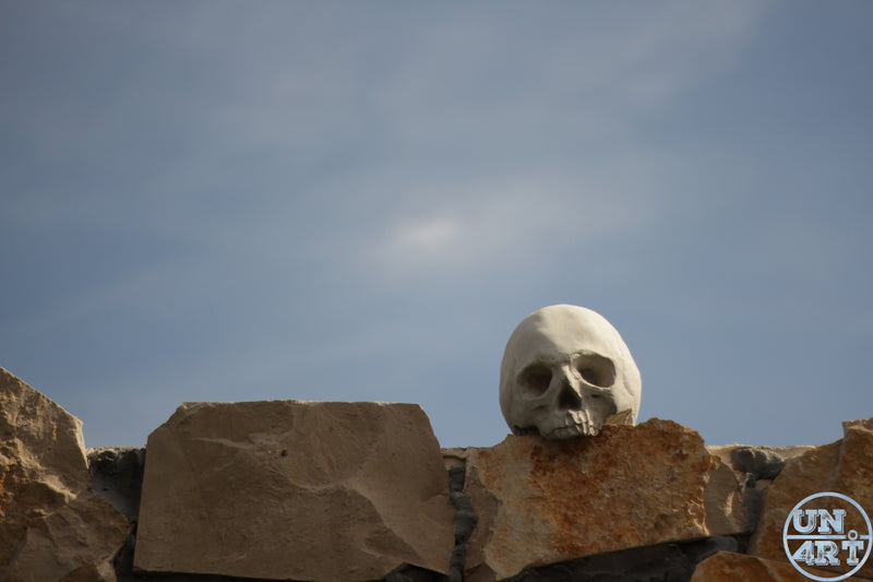 UN4RT imaginarium – pale white human skull resting on top of a rough dry stone wall under a bright hazy blue sky with the sun directly overhead, dark outdoor memento mori fine art photography.