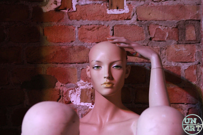 UN4RT imaginarium – bald female mannequin with painted green eyes and gold lips reclining with one hand raised to its head against a weathered red brick wall with peeling plaster in warm dim amber light, dark uncanny valley found-object fine art photography.