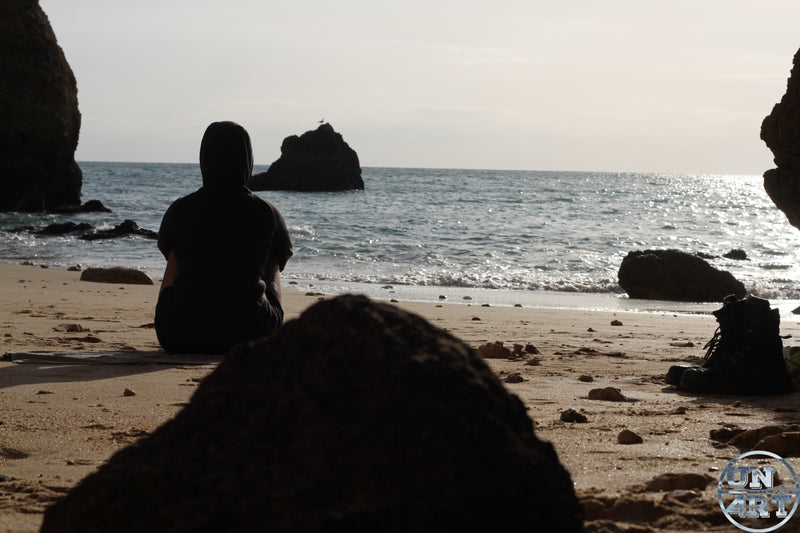 UN4RT imaginarium – rear silhouette of a solitary hooded figure seated on golden sand facing a calm silver-grey atlantic ocean with gentle waves and dramatic dark rock formations framing a secluded cove under a pale overcast sky with a pair of boots esting beside, dark contemplative solitude cinematic travel documentary photography.