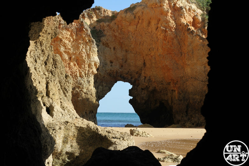 UN4RT imaginarium – view from inside a dark coastal cave looking outward through a large irregular natural rock arch opening revealing a smaller eroded rock arch beyond framing a bright teal Atlantic ocean horizon and a sunlit golden sandy beach with warm amber sandstone cliff walls under a clear blue sky, algarve coastal cave arch landscape documentary travel photography.