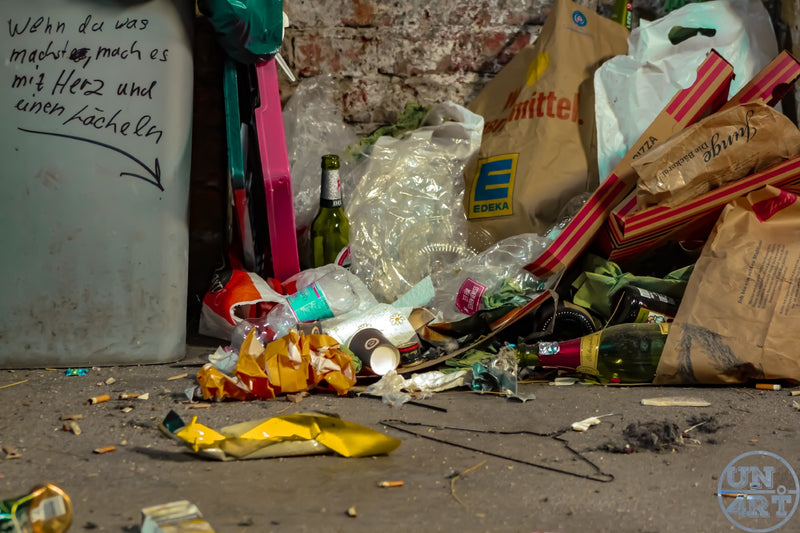 UN4RT imaginarium – close-up ground-level documentary photograph of a pile of urban street litter at night including crushed plastic bottles, glass beer and wine bottles, paper bags, food packaging, cigarette butts and assorted refuse scattered on a dirty pavement beside a grey refuse bin and a pink scooter against a weathered exposed brick wall with handwritten German graffiti reading "wenn du was machst, mach es mit herz und einem lächeln", dark urban waste social documentary street photography.