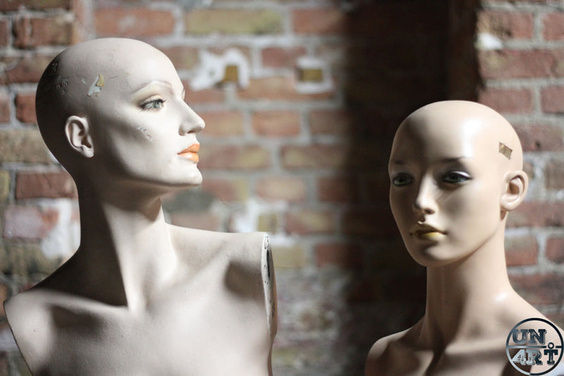 UN4RT imaginarium – close-up documentary photograph of two weathered vintage bald shop mannequin heads and busts with chipped matte beige skin, painted green and dark eyes and faded orange and gold lips posed side by side against a rough aged exposed red brick wall, one in three-quarter profile gazing upward left and one facing forward with a detached expression, dark found object identity uncanny valley street photography.