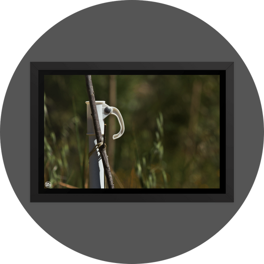 un4rt - the photographie shows a white umbrella stand with a wooden stick attached to it. the wooden stick is tied to the stand. very unsharp background with young trees and wild greenery.