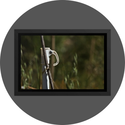 un4rt - the photographie shows a white umbrella stand with a wooden stick attached to it. the wooden stick is tied to the stand. very unsharp background with young trees and wild greenery.