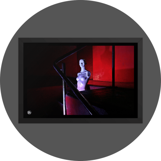 un4rt - the photographie shows the naked upper body of a mannequin without arms in the middle of a staircase. the mannequin is bold. the walls are all red, but due to the minimalistic lighting everything - except the mannequin - looks very dark. the point of view goes through a steel pipe handrail.