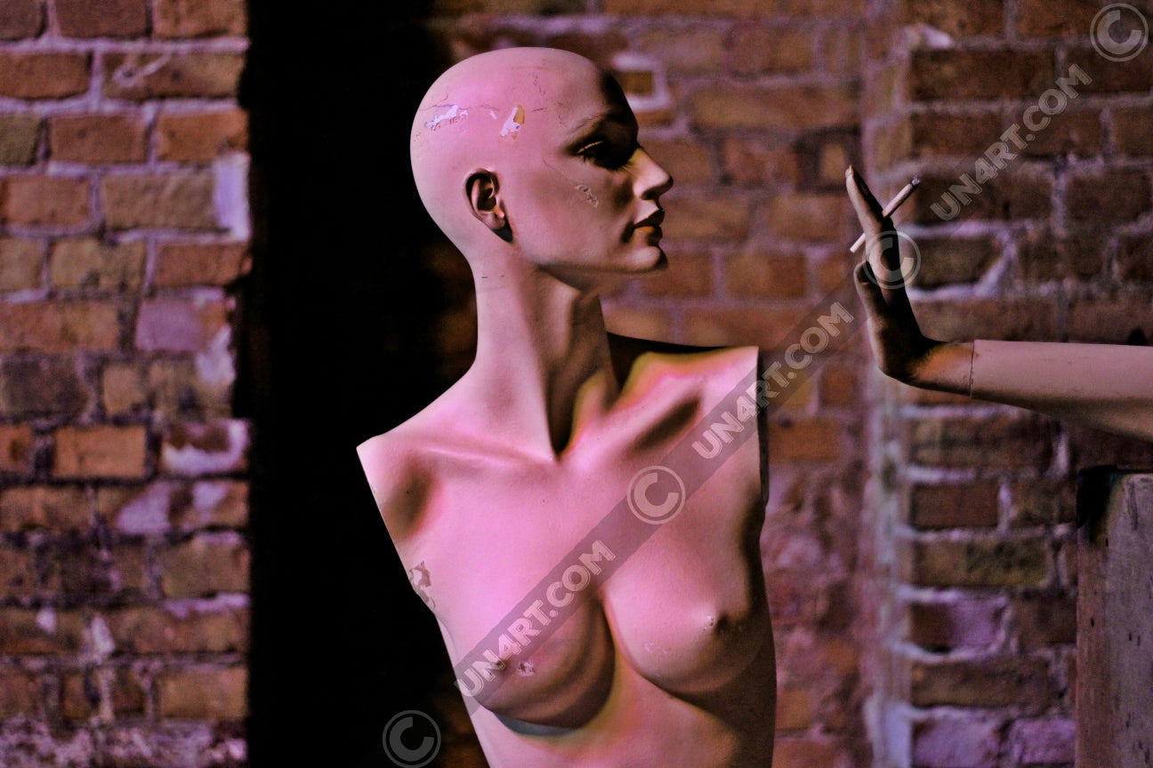 un4rt - the photographie shows the naked upper body of a bold mannequin without arms. the mannequin is looking sideways. a hand is offering a cigarette to the armless mannequin. brick stone wall in the background.