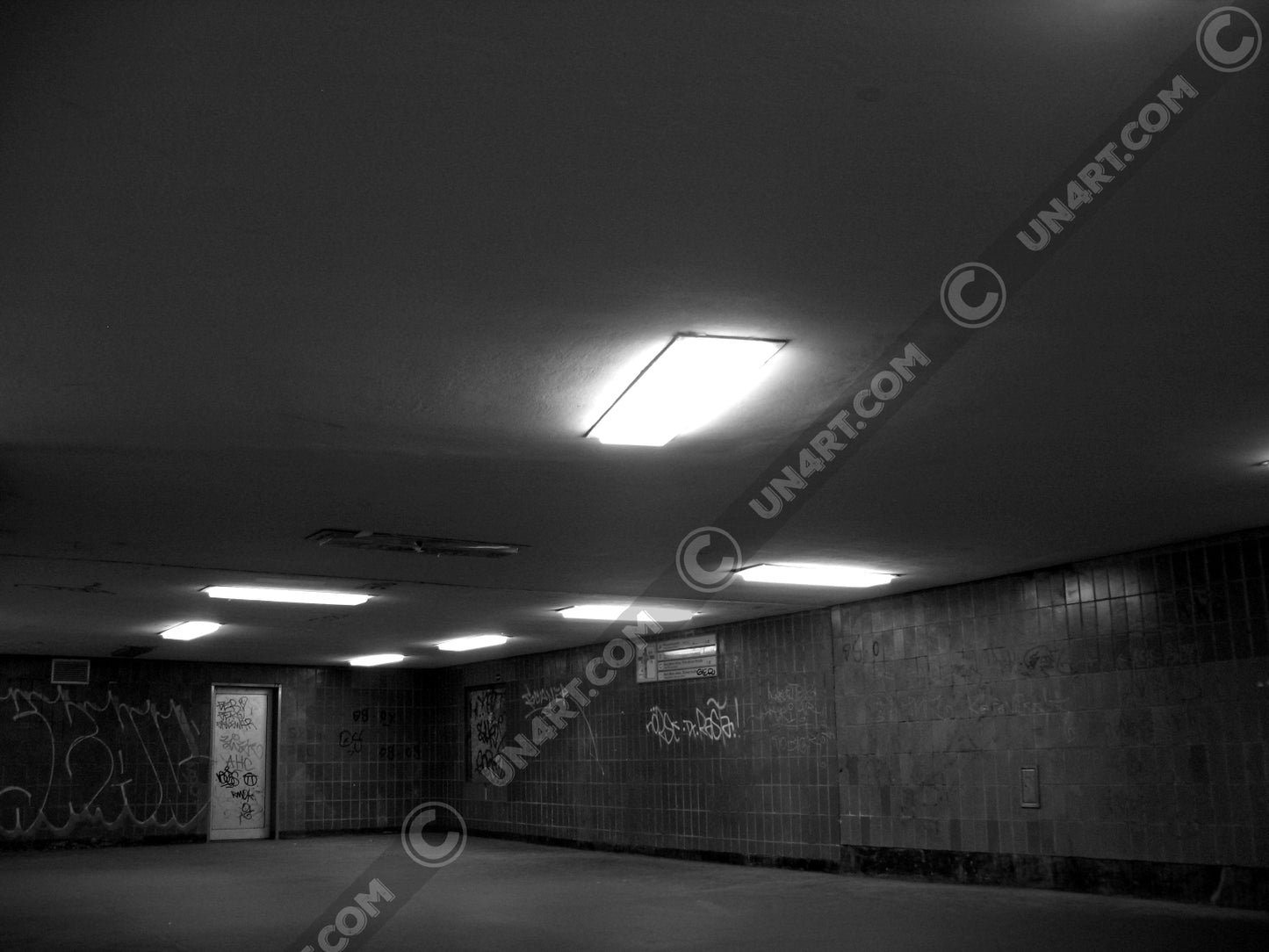 un4rt - a black and white photographie of a former underground trespas that once was part of the train station "alexanderplatz in berlin. the image shows a corner with a closed door. graffities on the wall. one lamp on the ceiling is broken.