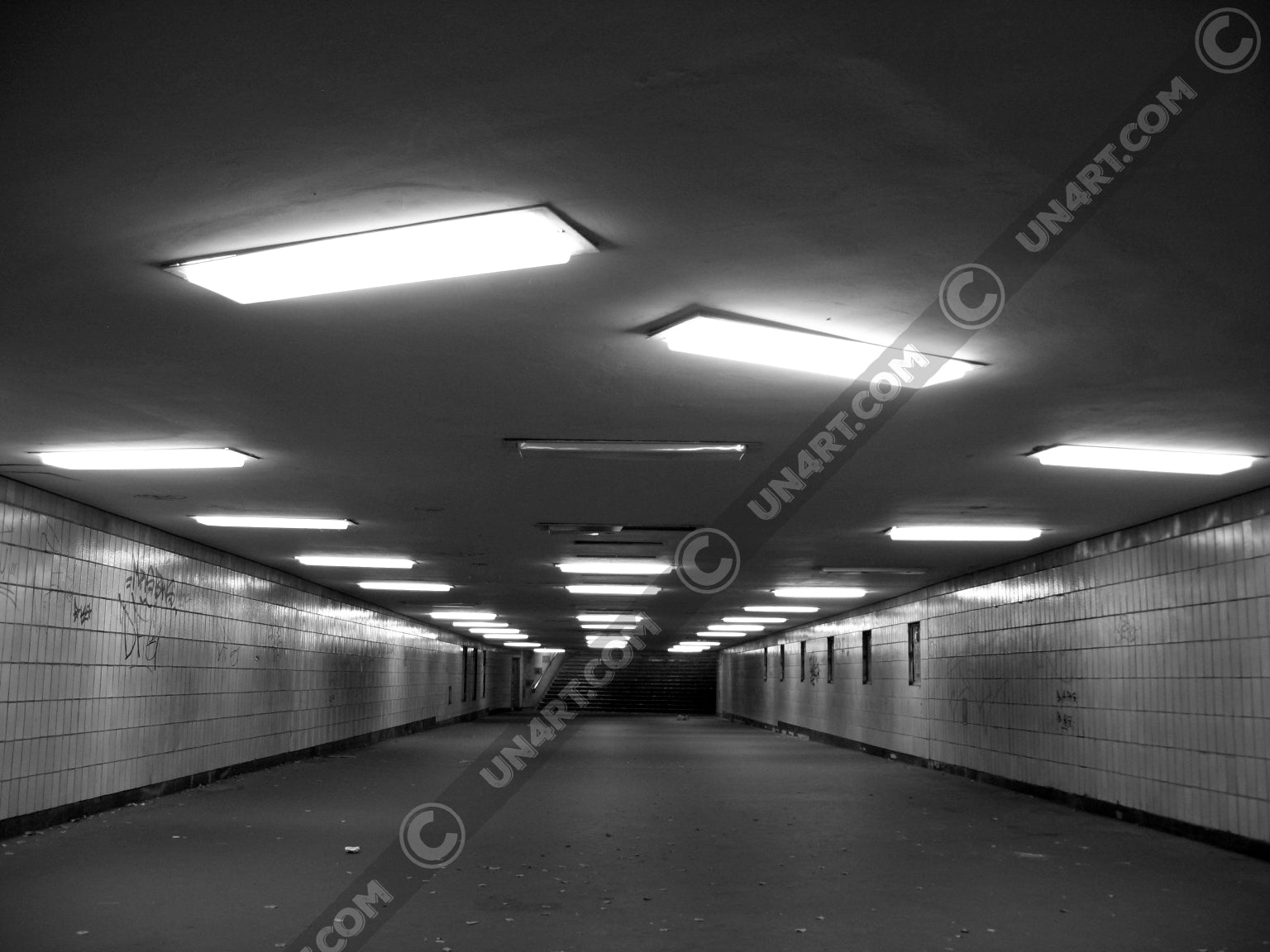 un4rt - a black and white photographie of a former underground trespas that once was part of the train station "alexanderplatz in berlin. the walls are tiled. some graffities on the walls and placeholders for advertising posters. trash on the floor. some lights n the ceiling don´t work.