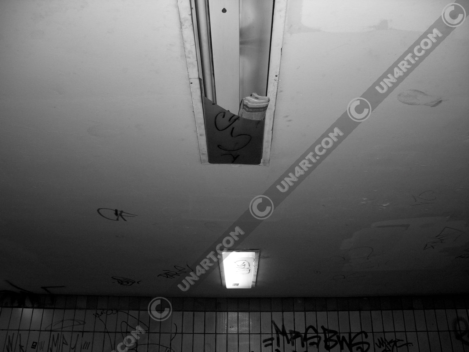 un4rt - a black and white photographie of a former underground trespas that once was part of the train station "alexanderplatz in berlin. the wall is tiled and coverd with graffities. one of the two lights on the ceiling don´t work anymore. the glas is broken and a newspaper is stuffed into the lamp.