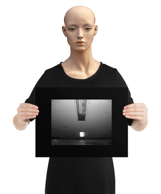 un4rt - a black and white photographie of a former underground trespas that once was part of the train station "alexanderplatz in berlin. the wall is tiled and coverd with graffities. one of the two lights on the ceiling don´t work anymore. the glas is broken and a newspaper is stuffed into the lamp. the image is held by a mannequin.