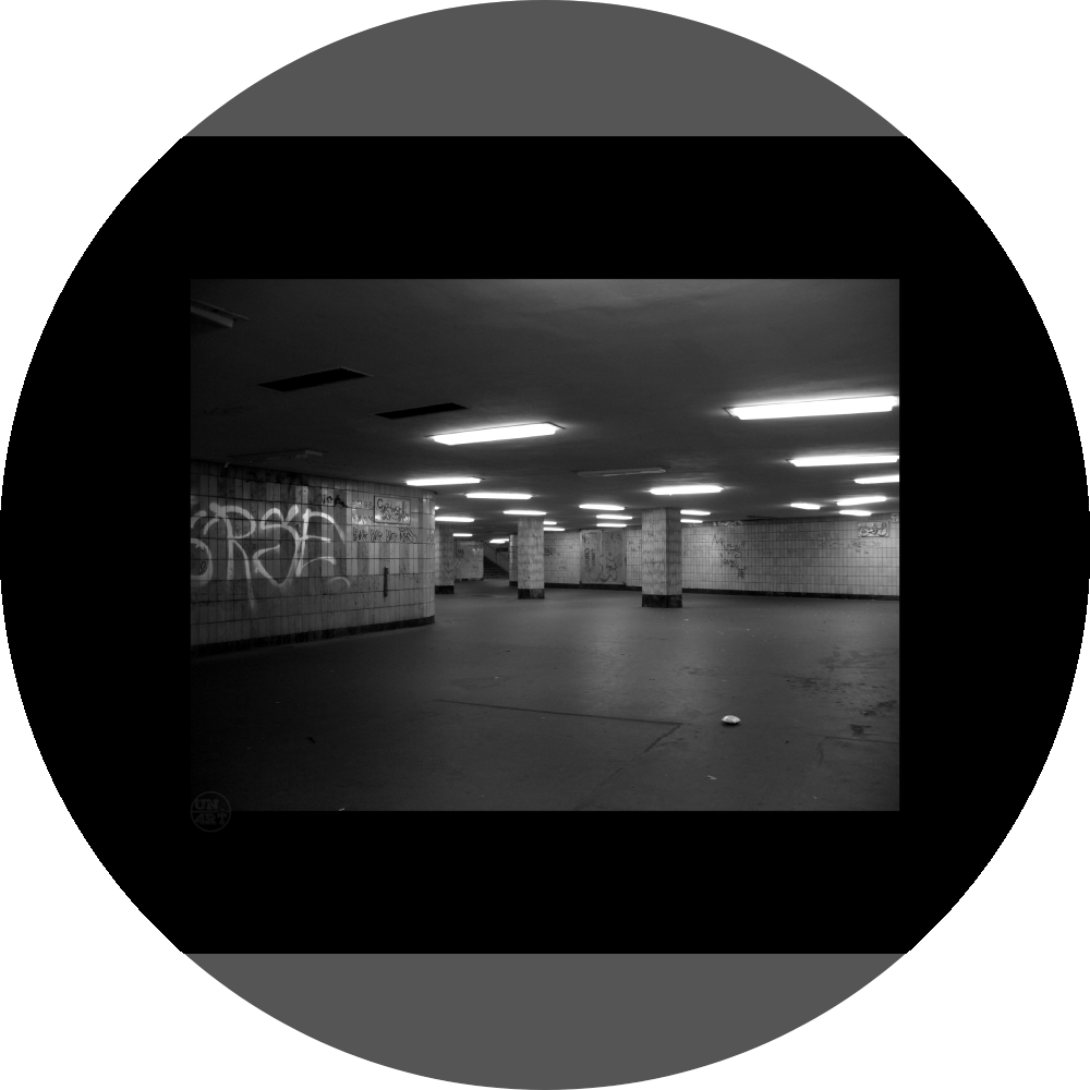 un4rt - a black and white photographie of a former underground trespas that once was part of the train station "alexanderplatz" in berlin. graffities on the wall, trash on the ground. some lights on the ceiling don´t work. a stairway at the end of the trespas.
