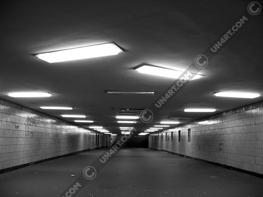 un4rt - a black and white photographie of a former underground trespas that once was part of the train station "alexanderplatz" in berlin. the walls are tiled. some graffities on the walls and placeholders for advertising posters. trash on the floor. some lights n the ceiling don´t work.