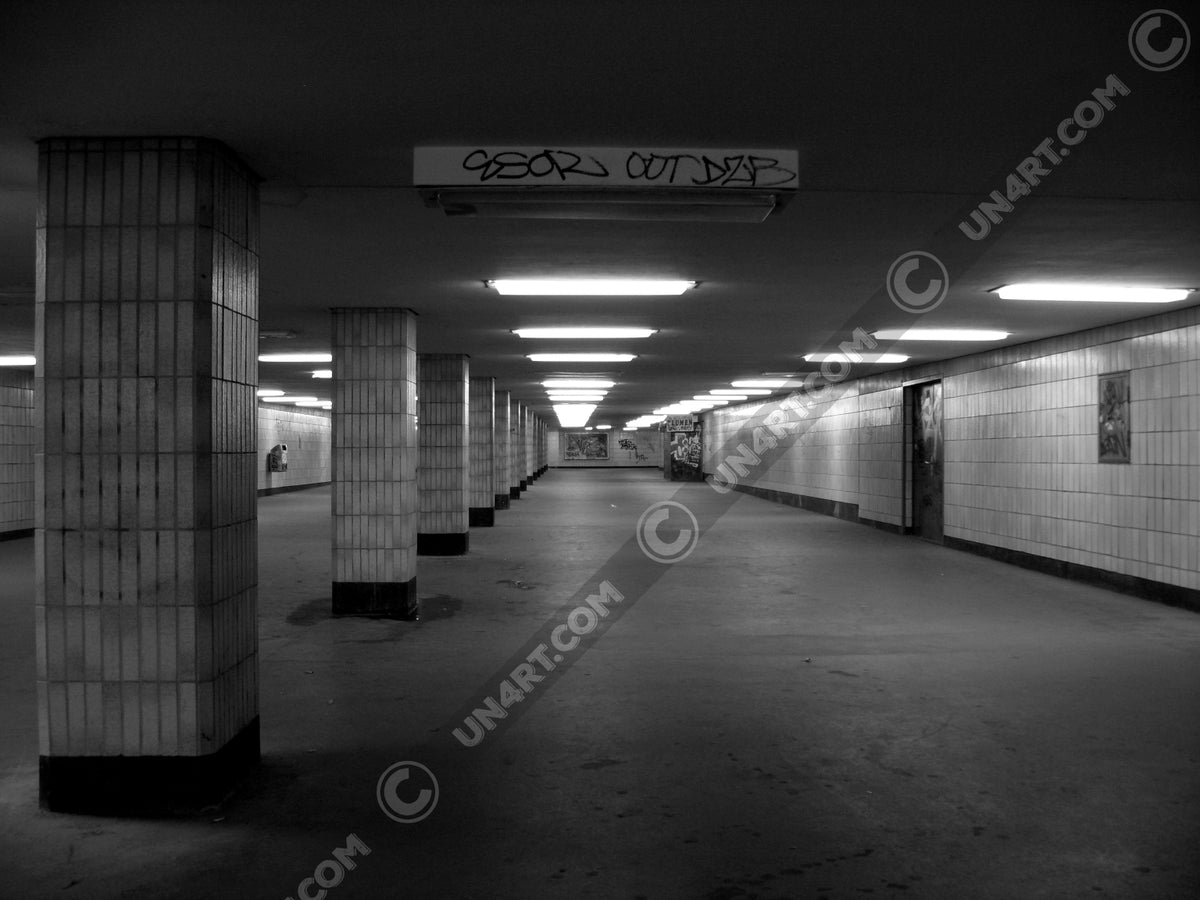 un4rt - a black and white photographie of a former underground trespas that once was part of the train station "alexanderplatz" in berlin. tiled square columns support the ceiling. a closed door on the right side. one light on the ceiling does´t work.