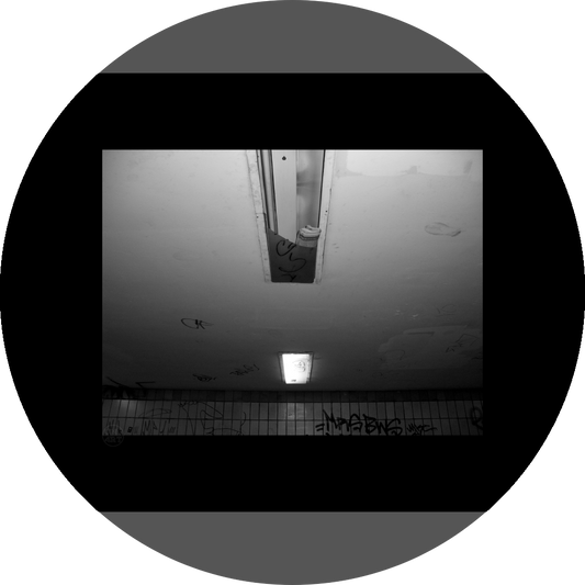 un4rt - a black and white photographie of a former underground trespas that once was part of the train station "alexanderplatz" in berlin. the wall is tiled and coverd with graffities. one of the two lights on the ceiling don´t work anymore. the glas is broken and a newspaper is stuffed into the lamp.