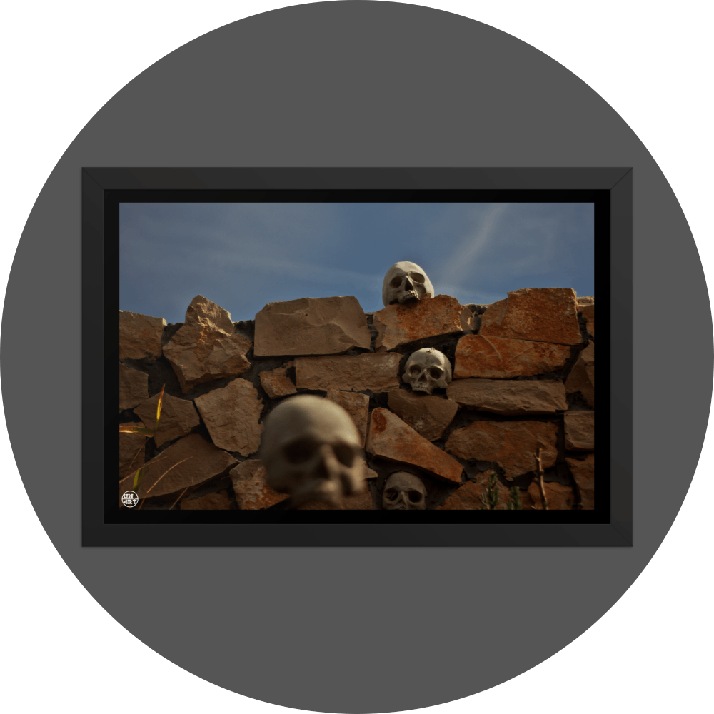 un4rt - the photographie shows a chaotic decorative stone wall with skulls in and on top of it. chemtrails in the sky starting to cover the natural blue of the sky.. one blurry skull in the foregrund.